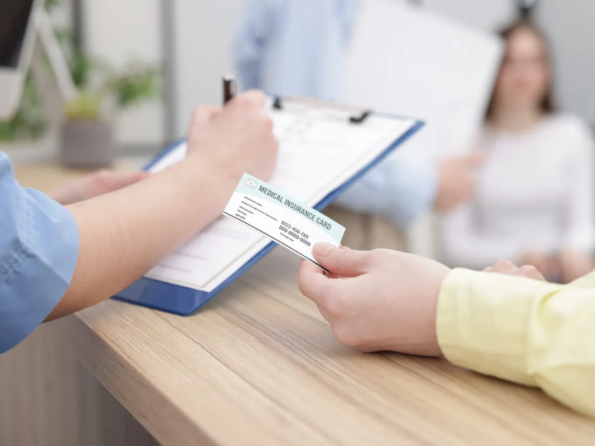 Stock photograph of models: a patient hands a health insurance card across a clinic counter to a receptionist holding a clipboard.