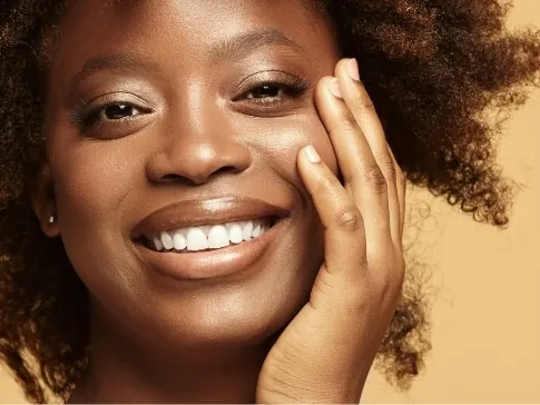 Stock photograph of a model: smiling woman with curly hair resting her hand against her cheek on a tan background.