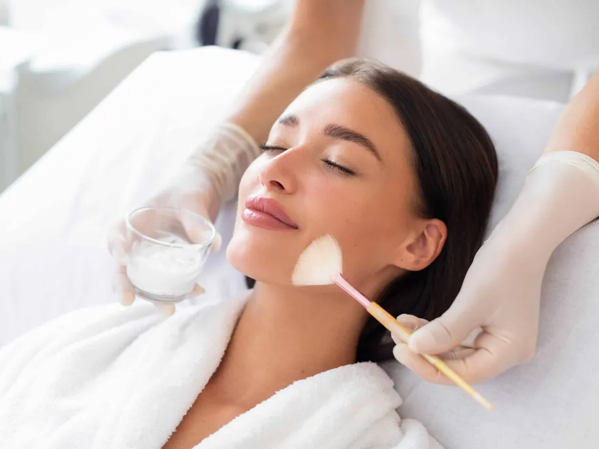 Stock photograph of a model: a woman in a white robe lies back with her eyes closed while a gloved hand brushes a peel onto her face
