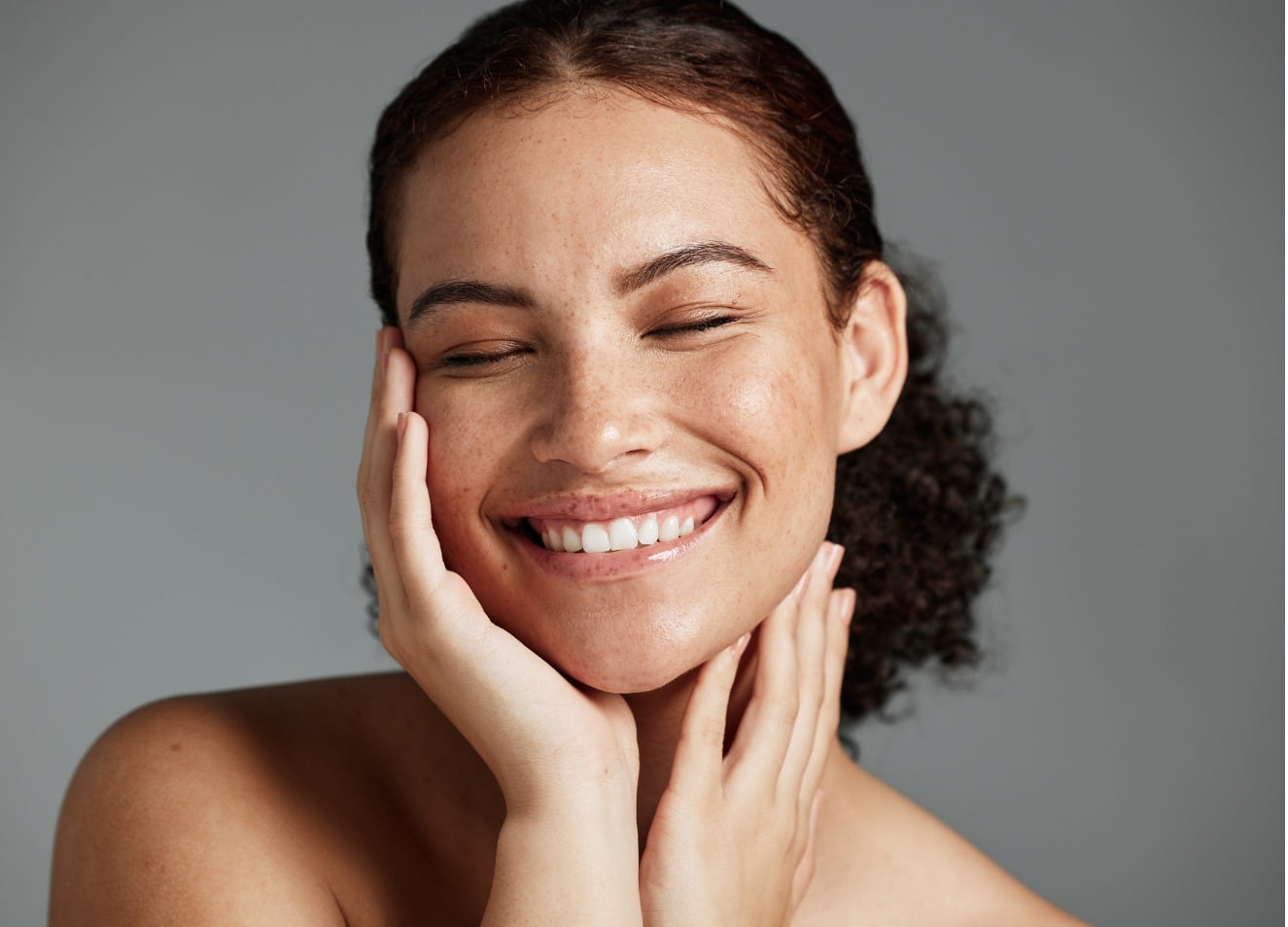 Stock photograph of a model: a smiling woman with freckles and curly hair, eyes closed, rests her fingertips on her cheek against a grey backdrop