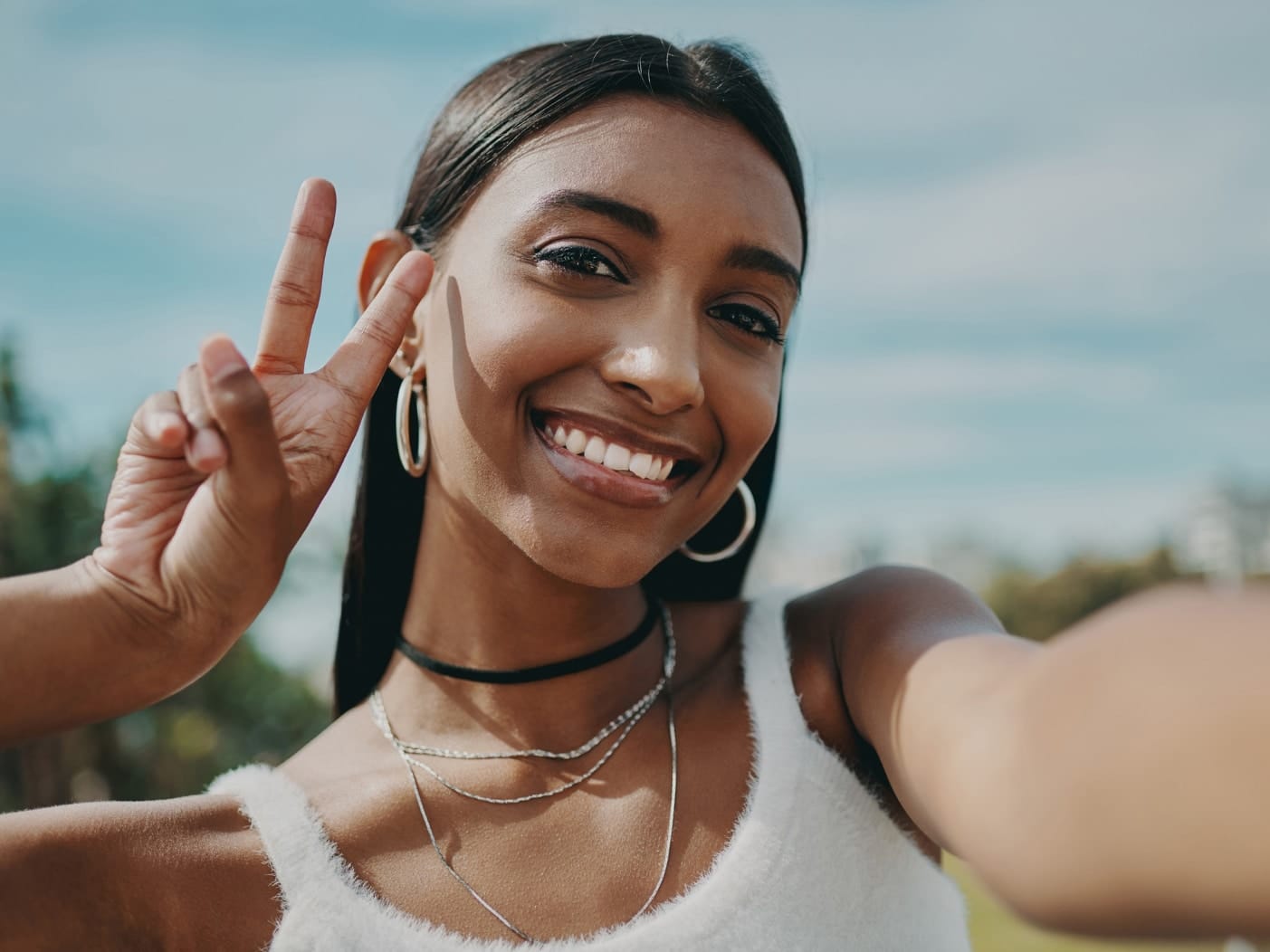 Stock photograph of a model: a smiling woman with dark hair and hoop earrings makes a peace sign while taking a selfie outdoors