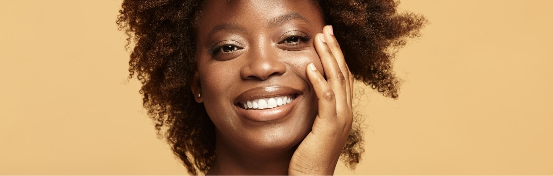 Stock photograph of a model: smiling woman with curly hair resting her hand against her cheek on a tan background.