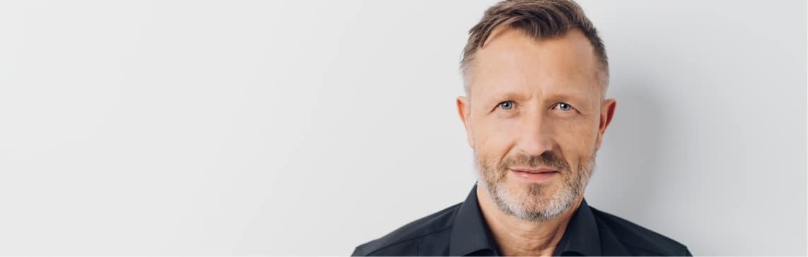 Stock photograph of a model: man with short graying hair and a beard looking at the camera in a dark shirt.