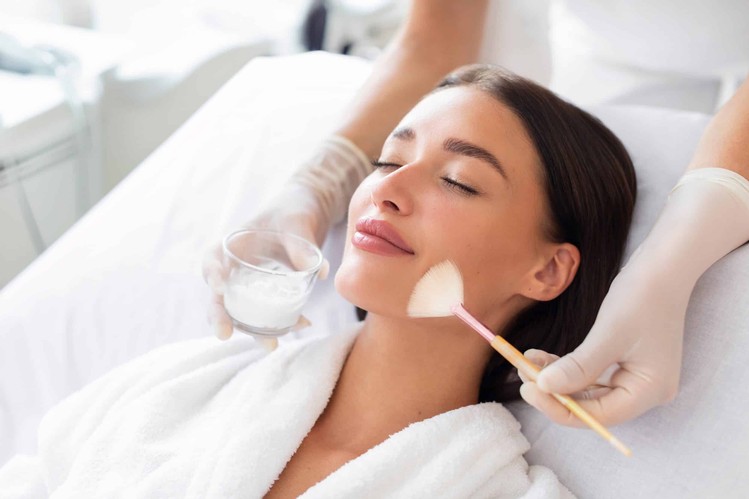 Stock photograph of a model: a woman in a white robe lies back with her eyes closed while a gloved hand brushes a peel onto her face