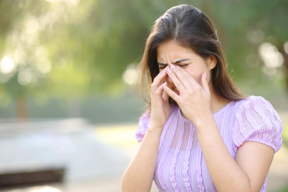Stock photograph of a model: a woman in a lilac blouse rubs her eyes and nose outdoors, as if from a spring allergy